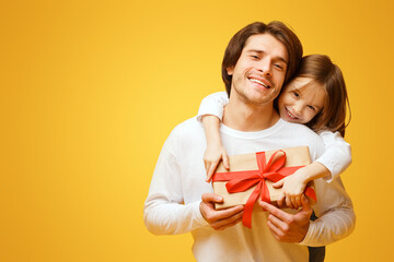 Wall Mural - Cute little daughter and father holding present, girl hugging dad from behind, Father Day concept, yellow background