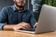 © Rutchaneewan - Focused programmer engaged in coding session, modern workspace with laptop, displaying lines of code, denim shirt, productive atmosphere, creative tech environment.