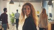 © Riko - Smiling young woman in business attire at modern office with colleagues in the background.