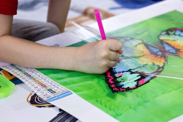 A boy collecting diamond embroidery. Selective focus. Close-up of a boy's hand assembling a multicolored diamond mosaic butterfly.