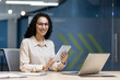 © Liubomir - Businesswoman with glasses holding digital tablet in office. Smiling professional using technology at desk, conveying confidence and efficiency. Modern workspace equipped with laptop and notepad