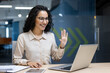 © Liubomir - Businesswoman in office using laptop for virtual meeting, smiling and waving hand. Engaging in professional communication, fostering connection, and showcasing confidence using technology.