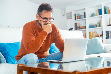 Concentrated adult male using his laptop on the sofa at home, navigating an alternative office setting while managing his small online business or planning for the next vacation getaway