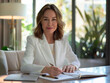 © Jirawatfoto - professional woman in white blazer sits at desk, writing in notebook with confident smile. modern office setting features plants and natural light, creating inspiring atmosphere