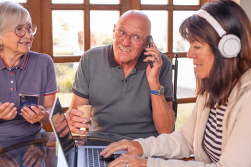 Wall Mural - Happy group of caucasian seniors using laptop sitting at table against window at home. Three modern old generation pensioners and new technologies