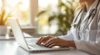 © Anastasiia Havelia - A close-up photo of a female doctor typing on a laptop computer, with a light background and a blurred office in the background, creating a bokeh effect. The doctor is wearing a wh