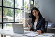 © apichat - A woman is sitting at a desk with a laptop and a cell phone