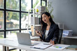 © apichat - A woman is sitting at a desk with a laptop and a cell phone