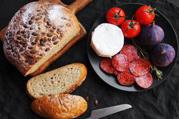 Plate with appetizers of Brie cheese, perroni, tomatoes, figs and rosemary sprigs next to sliced ​​grain bread on a dark background