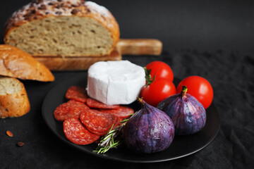 Plate with appetizers of Brie cheese, perroni, tomatoes, figs and rosemary sprigs next to sliced ​​grain bread on a dark background