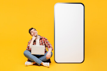 Wall Mural - Full body young IT man he wears red checkered shirt casual clothes big huge blank screen mobile cell phone smartphone with area hold use work on laptop pc computer isolated on plain yellow background.