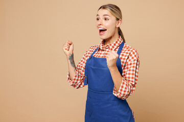 Wall Mural - Young woman barista bartender barman employee wear blue apron shirt work in coffee shop do winner gesture celebrate clenching fists isolated on plain beige background. Small business startup concept.