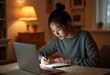 © Jittiwan - A young lady writing note in a paper with a laptop at table in a house at night scene
