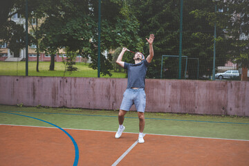 Badminton player focuses on the shuttlecock mid-air during a match on an outdoor court. With his racket raised, he prepares for the next shot, surrounded by an urban and scenic background