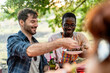 © Lomb - Diverse friends sharing food at outdoor picnic, multicultural men enjoying meal in nature