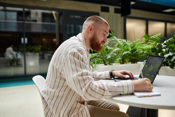 Side view of concentrated adult man using laptop and writing in notebook while studying or working at table in office center cafe, copy space