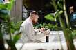 © Seventyfour - Shot through green living plants of concentrated businessman using digital tablet to work or study remotely sitting at table in cozy sunlit cafe in contemporary office center, copy space