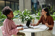 © Seventyfour - Side view of African American woman discussing work strategy with female colleague while working on laptop at cafe table in contemporary business center, copy space