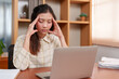 © Ekkasit A Siam - Fatigued office employee experiences severe migraine, fingers on forehead, in front of a laptop, at home workspace. Work-related stress visible on person's face, seeking relief from head pain