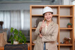© Ekkasit A Siam - Asian female architect wearing white hard hat sitting at desk using tablet smiling in office wearing beige blazer working on project design modern work environment.