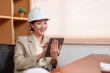 © Ekkasit A Siam - Asian female architect smiling standing in office holding white hard hat rolled blueprint papers wearing beige blazer bookshelf background professional worker in work environment.