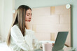 © Ekkasit A Siam - Asian woman working from home feeling tired resting chin on hand in front of laptop wearing white blouse focused on screen working from bedroom home office environment.