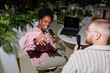 © Seventyfour - Cheerful female podcast host of African American Ethnicity playfully waving hand towards camera captured recording new episode with male guest at table in cafe with green plants, camera flash