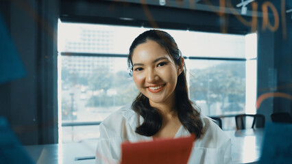 Beautiful female leader thinking about start up project in front of glass board. Smiling young project manager looking at colorful sticky notes with show creativity marketing strategy. Manipulator.