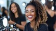 © Pinklife - Individual with curly hair seated in a makeup studio, wearing a black outfit, surrounded by cosmetics and other people in the background involved in makeup activities.