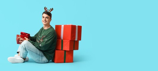 Wall Mural - Happy young man in Christmas sweater and with gift boxes on light blue background with space for text