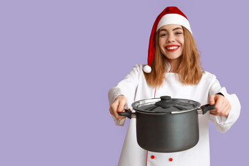 Canvas Print - Young female chef in Santa hat with cooking pot on purple background