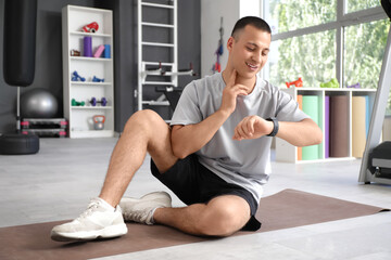 Wall Mural - Sporty young man with smartwatch checking pulse on mat in gym