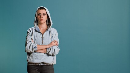 Wall Mural - Bold serious woman putting her hood on and acting tough in studio, posing with arms crossed and showing confidence in her street style. Modern independent person acts as a gangster. Camera B.