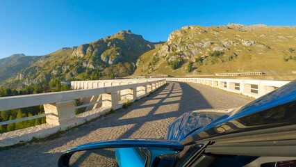 CLOSE UP: Picturesque shot of the Italian mountains ahead of the luxury car driving along an old road. Metallic blue car cruises along the cobblestone bridge overlooking the breathtaking Dolomites.