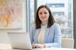 © Tada - usiness woman sitting at a desk with a laptop, looking directly into the camera, wearing a light blue suit and a white blouse, with her brown hair in a straight, long bob cut hairstyle