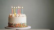 © Shisanupong - A white frosted birthday cake with colorful sprinkles and lit candles, placed on a marble cake stand against a neutral background.