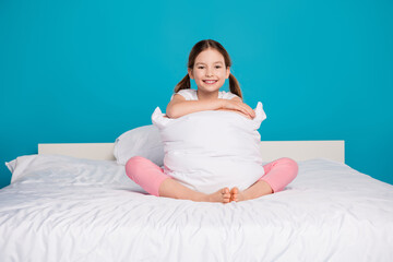 Poster - Full length photo of adorable little girl hugging pillow sit bed sleepover bedroom dressed stylish pajama isolated on blue color background