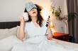 © _KUBE_ - Portrait of funny sleeping young woman wearing sleep mask call with cup and drink with phone while sitting in bed. Concept of being late for work and morning awakening
