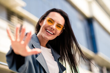 Wall Mural - Photo of cheerful pretty lady dressed grey jacket dark eyewear waving arm hi enjoying sunshine outdoors town street