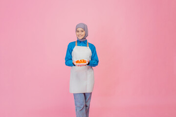 A cheerful woman in a cozy apron presents fresh vegetables against a vibrant pink backdrop