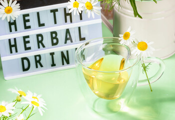 Wall Mural - Herbal medicine. Healthy chamomile tea in glass cup, flowers, inscription by black letters on green.