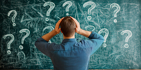 Fototapete - Man holding his head in front of a chalkboard filled with question marks in a classroom..