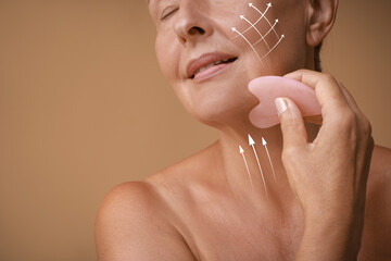 Sticker - Woman doing facial massage with gua sha tool on pale brown background, closeup. Arrows on skin showing direction of moves