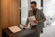© Moon Safari - Portrait of smiling male printer in eyeglasses holding documents and operating computer printer in office