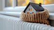 © ERiK - Closeup of a small model house wrapped in a warm woolen scarf above a white radiator. Cold protection, home insulation, space heating system, energy crisis concept.
