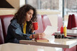 © pressmaster - Portrait of young man sipping soda in red cup while sitting in diner booth with camera and condiments on table, exuding relaxed vibe and long curly hair