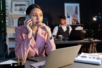 Focusing woman calling to customer working at front desk view while waiting email business project report on laptop at late night time with blurry man coworker background at modern office. Postulate.