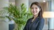 © EarthWalker - Confident female business leader in black suit looking into camera in modern office with a green indoor plant. Portrait of beautiful smiling Asian businesswoman