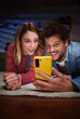 © CarlosBarquero - Vertical young Caucasian couple looking surprised face at phone lying on carpet in living room at night home. Millennial relationship enjoying mobile together. Domestic people leisure and technologies