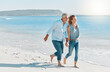 © KayExam/peopleimages.com - Mature couple, running and holding hands with beach for holiday, weekend or outdoor bonding. Man, woman or lovers with smile for vacation, stroll or view in sunshine on sandy shore by ocean coast